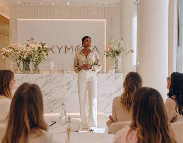 A refined woman standing at the front of a modern luxury beauty training room, presenting to a group of elegant female students. She wears a silk blouse and wide-leg trousers in neutral tones, with natural makeup and pulled-back hair. The room is spacious and filled with soft daylight, beige and white tones, minimalistic yet sophisticated. Marble tables, whiteboard or projection screen behind her, subtle branding. Elegant, calm atmosphere, MAXYMOVA training vibe.